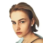 Український Альянс Жінка з коротким світлим волоссям, у перлинній сережці та світло-блакитному топі, дивиться прямо в камеру на білому тлі.