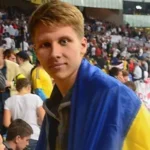 Молода людина, загорнута в синьо-жовтий прапор, стоїть на переповненому стадіоні й дивиться в камеру. Люди на задньому плані, судячи з усього, спостерігають за якимось заходом. ГО Український Альянс